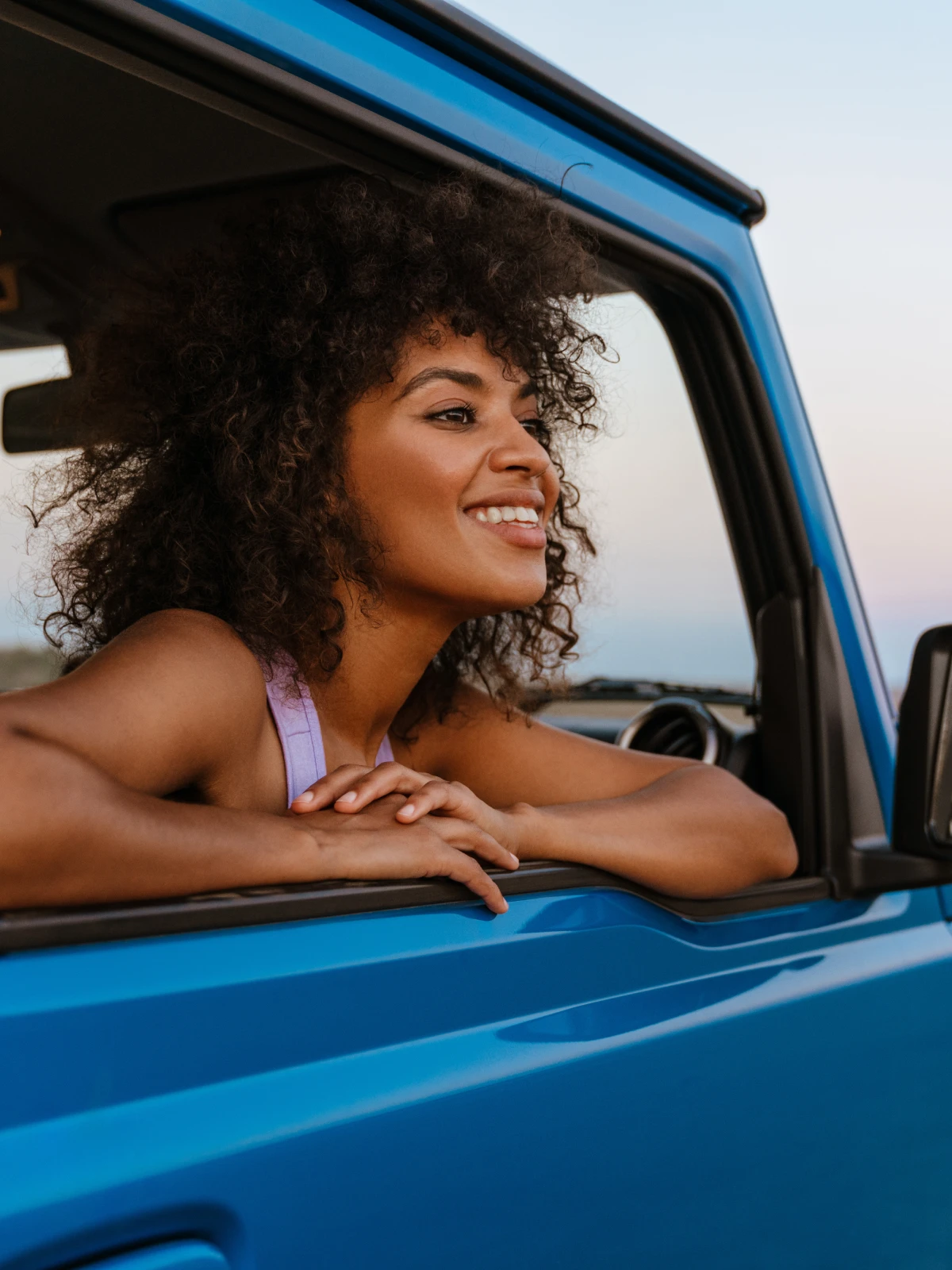 Woman smiling and waving from a red convertible on a coastal road at sunset