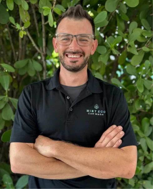 Guy Kean, Mint Eco Car Wash team member, smiling in a black Mint Eco Car Wash polo shirt in front of green foliage